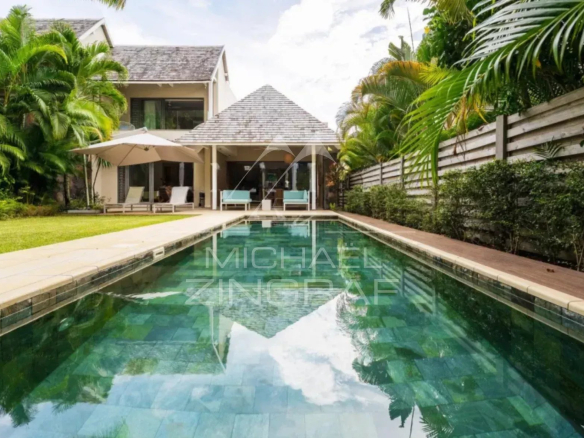 Villa-style backyard with a rectangular pool, lounge chairs under a large umbrella, and a shaded seating area at the house patio surrounded by tropical plants and a wooden fence.