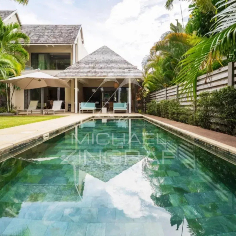 Villa-style backyard with a rectangular pool, lounge chairs under a large umbrella, and a shaded seating area at the house patio surrounded by tropical plants and a wooden fence.