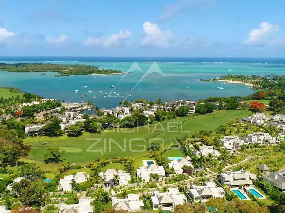 Aerial view of a coastal resort with white villas, palm trees, a golf course, and turquoise lagoons with boats near the shoreline.