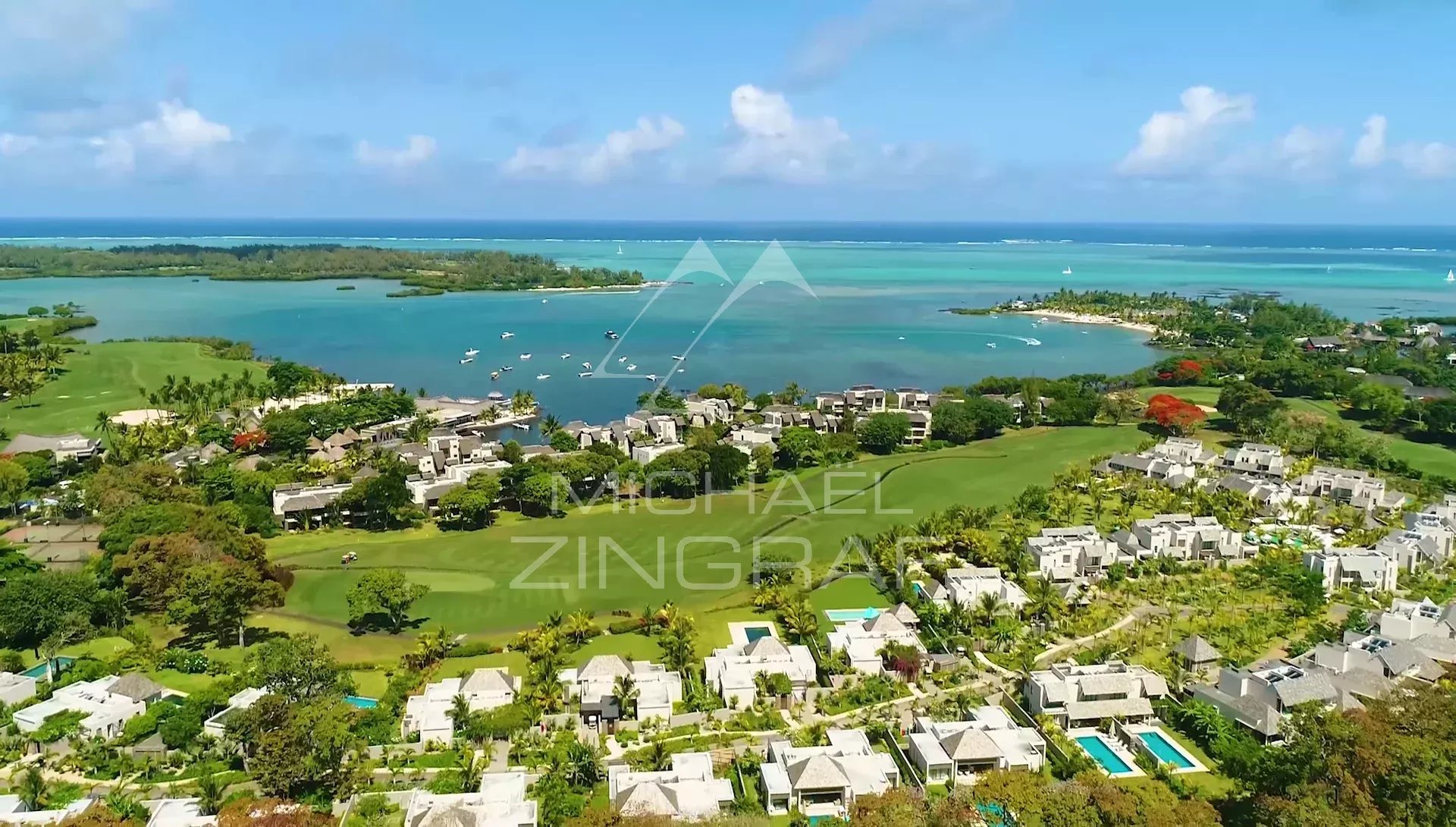 Aerial view of a coastal resort with white villas, palm trees, a golf course, and turquoise lagoons with boats near the shoreline.