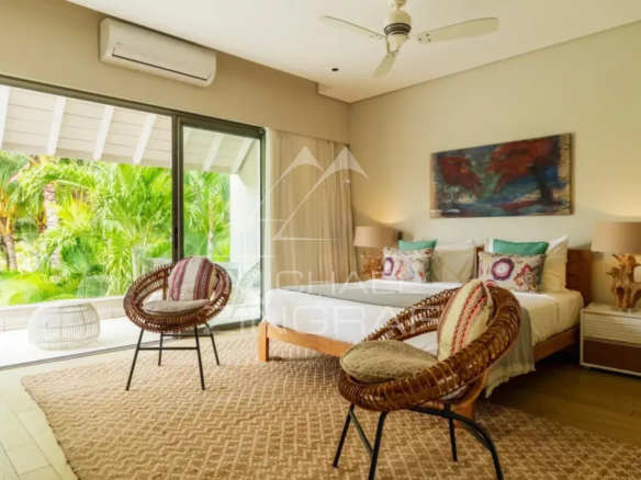 Bright hotel room with two single beds, colorful pillows, and wicker chairs facing a glass sliding door to a tropical patio garden outside.