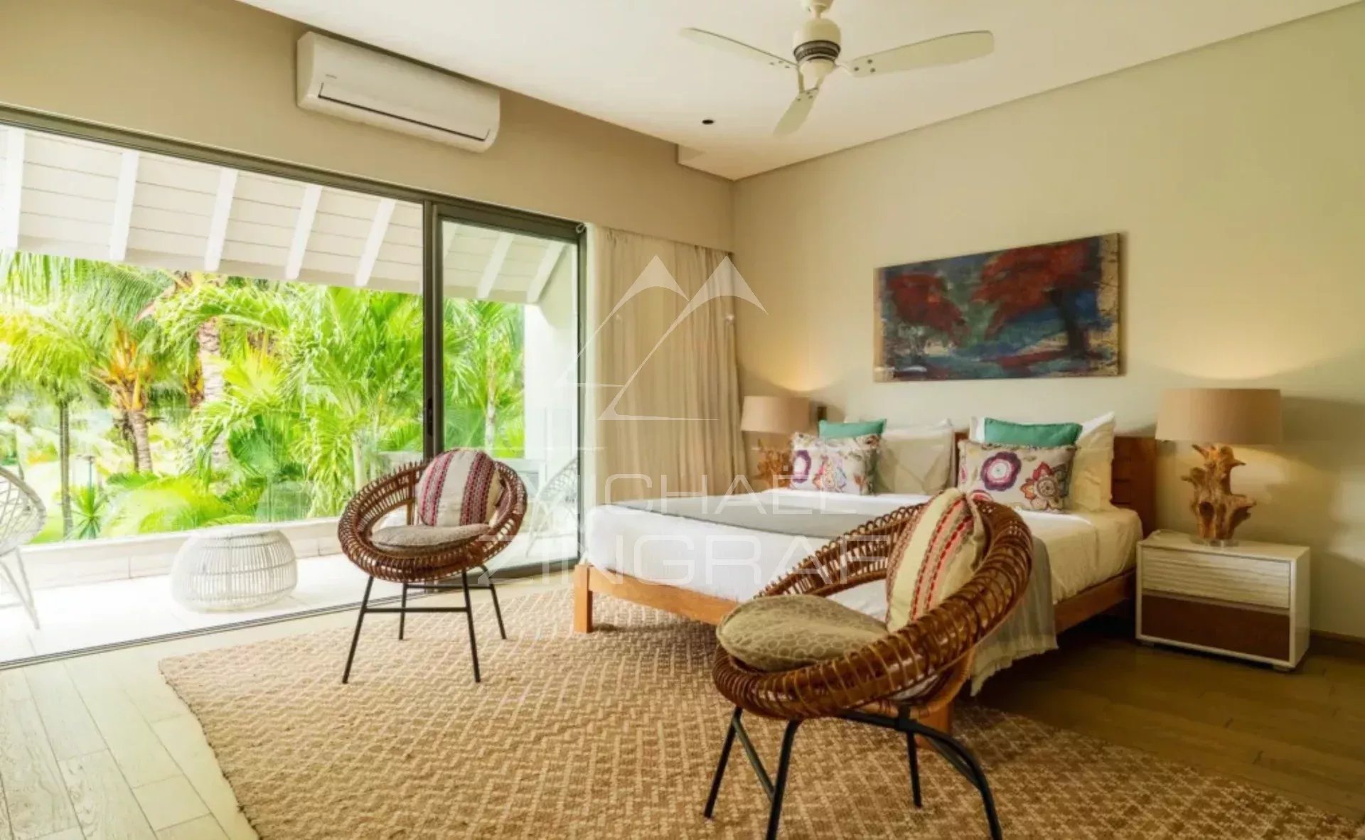 Bright hotel room with two single beds, colorful pillows, and wicker chairs facing a glass sliding door to a tropical patio garden outside.