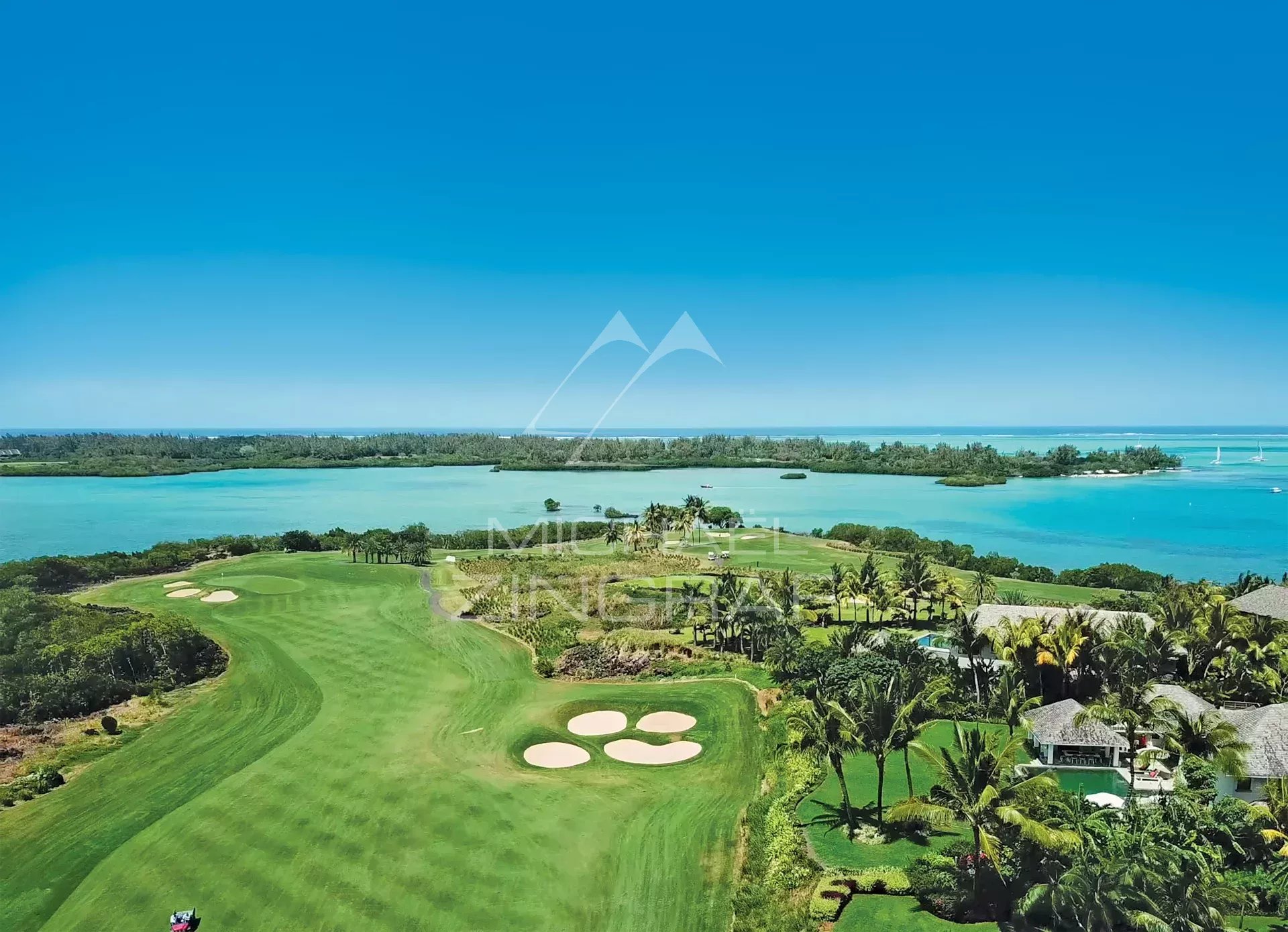 Aerial view of a coastal golf course with turquoise water, green fairways, and palm trees nearby.