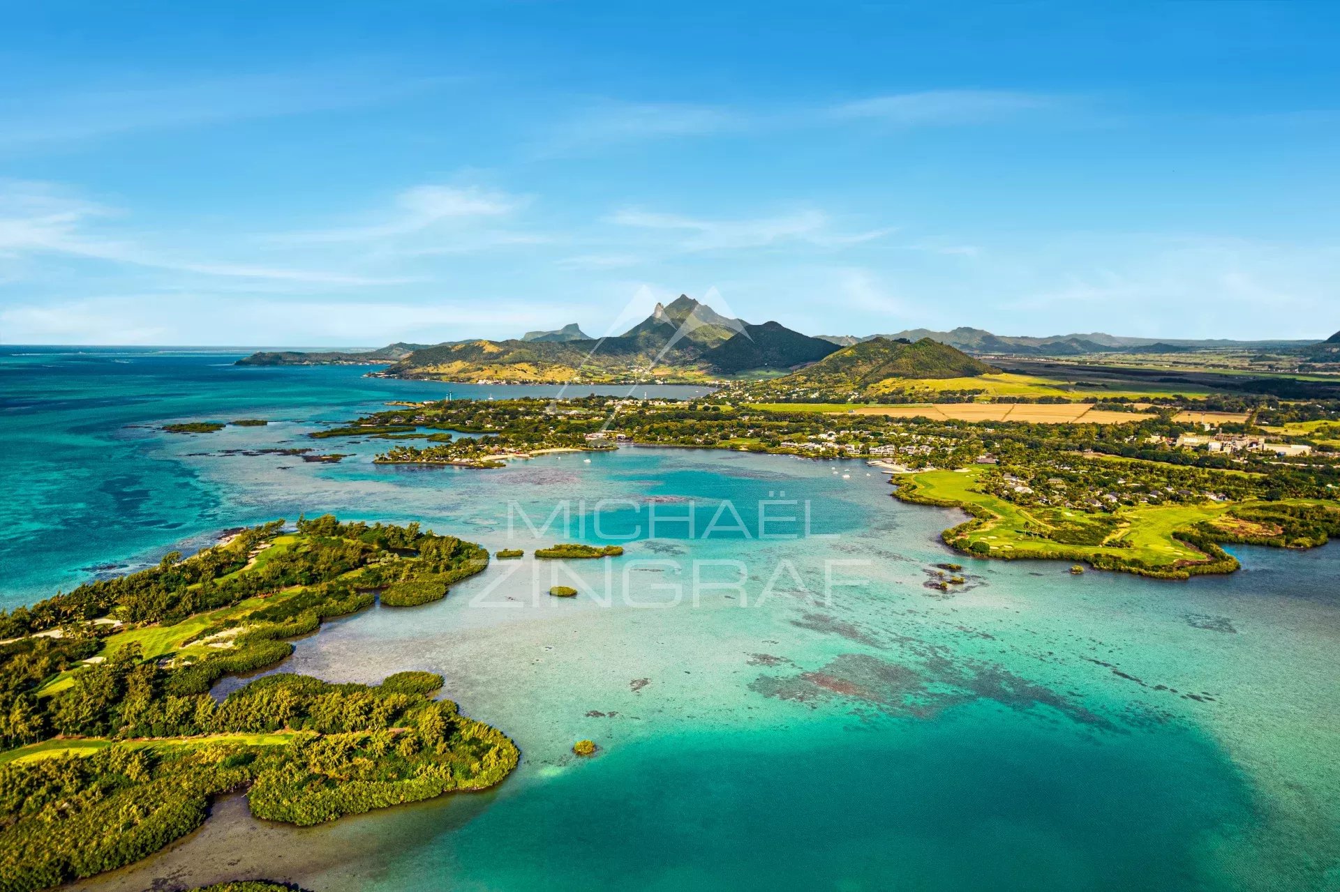 Aerial view of a tropical coastline with turquoise lagoons, green islands, and a small village along the shore, with mountains in the distance.