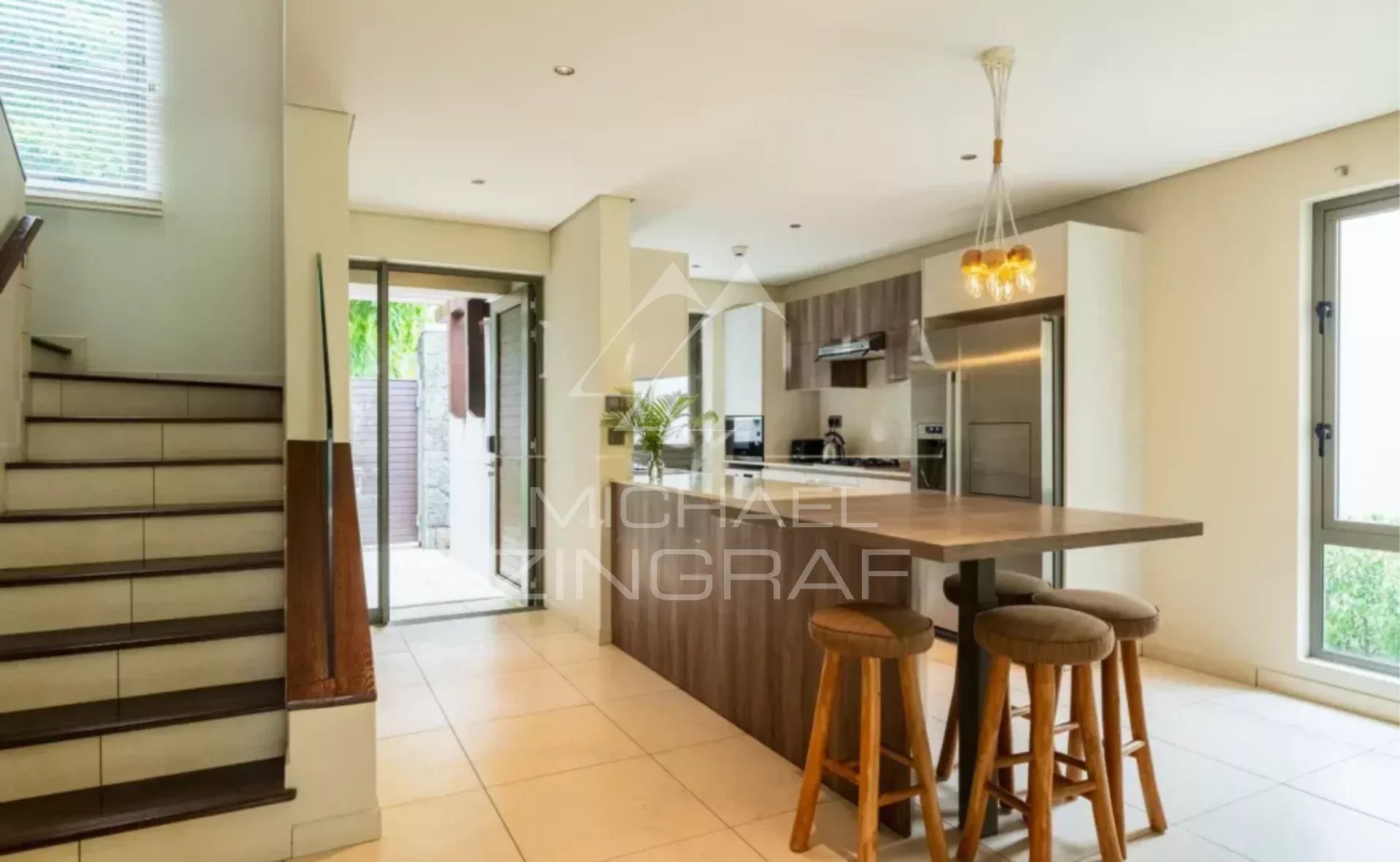 Open-plan kitchen with a wooden island and three bar stools, stairs to the left, glass doors to outdoors.