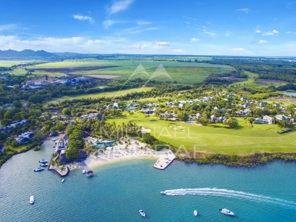 Aerial view of a tropical waterfront resort: white buildings surround a marina, sandy beach, and calm turquoise bay with several boats. Green lawns and golf fairways extend inland under a blue sky.