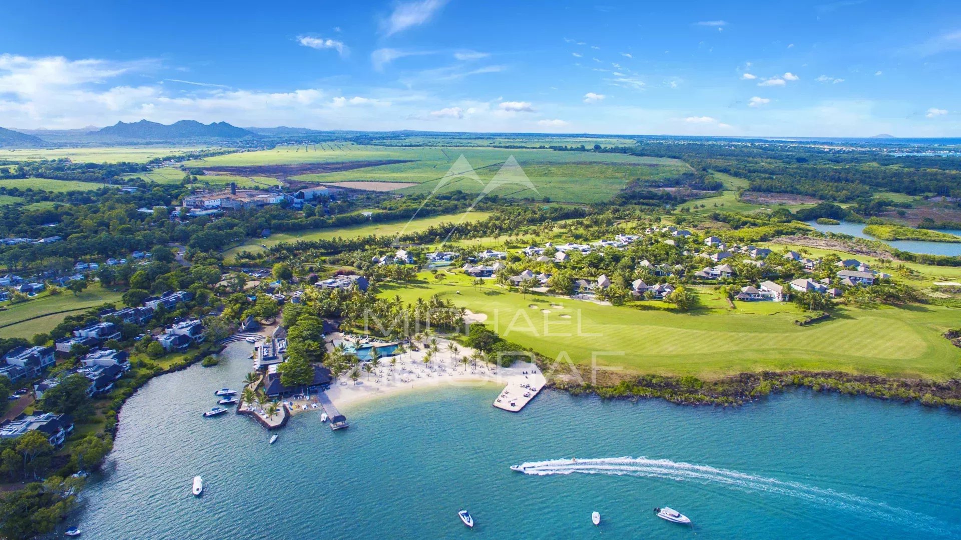 Aerial view of a tropical waterfront resort: white buildings surround a marina, sandy beach, and calm turquoise bay with several boats. Green lawns and golf fairways extend inland under a blue sky.
