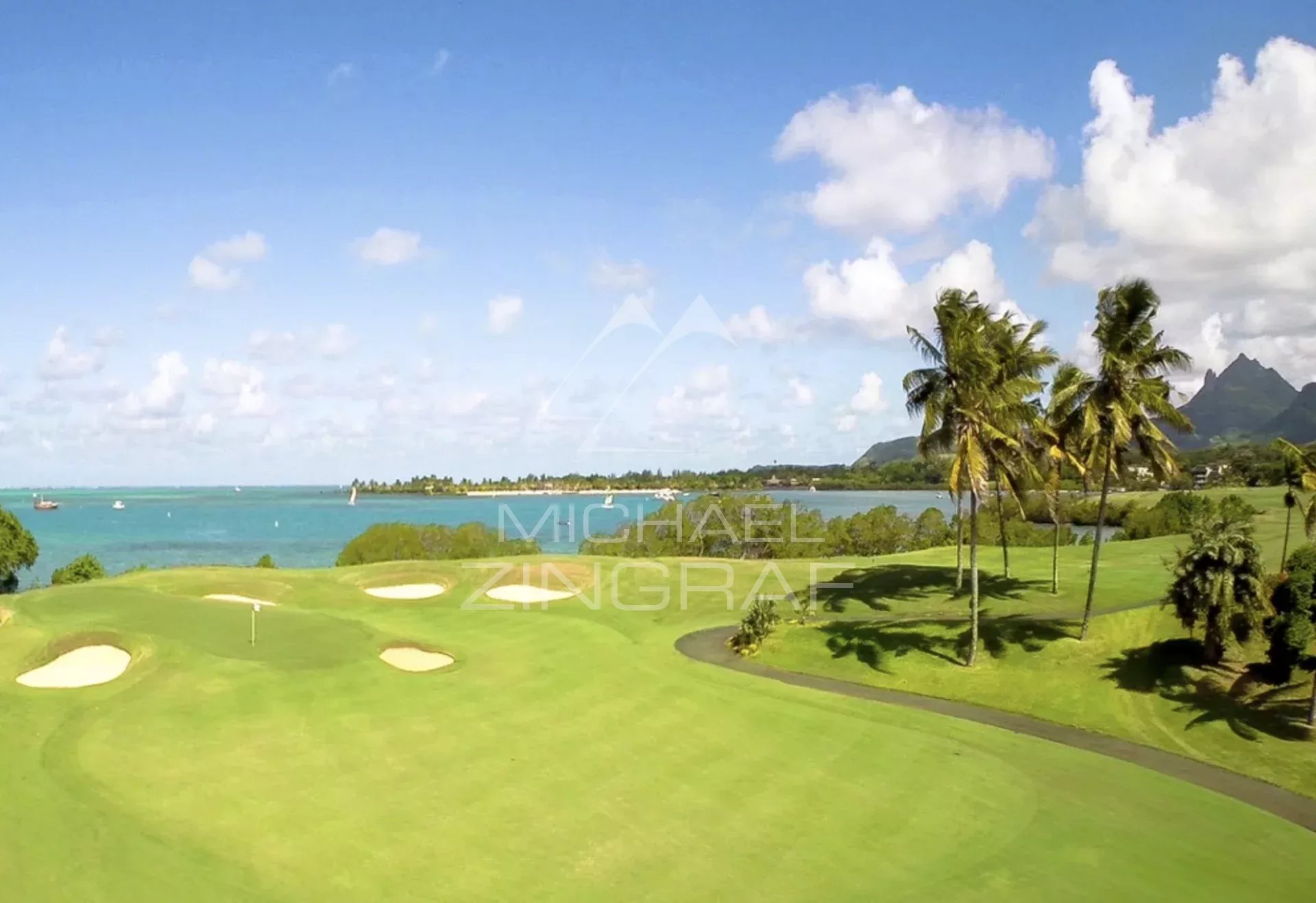 Coastal golf course with green fairways, sand bunkers, and palm trees along a turquoise sea under a bright blue sky.