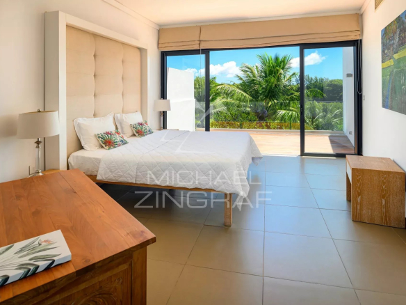Hotel bedroom with a king bed, tufted headboard, nightstands, and sliding glass doors to a balcony overlooking palm trees and blue sky.