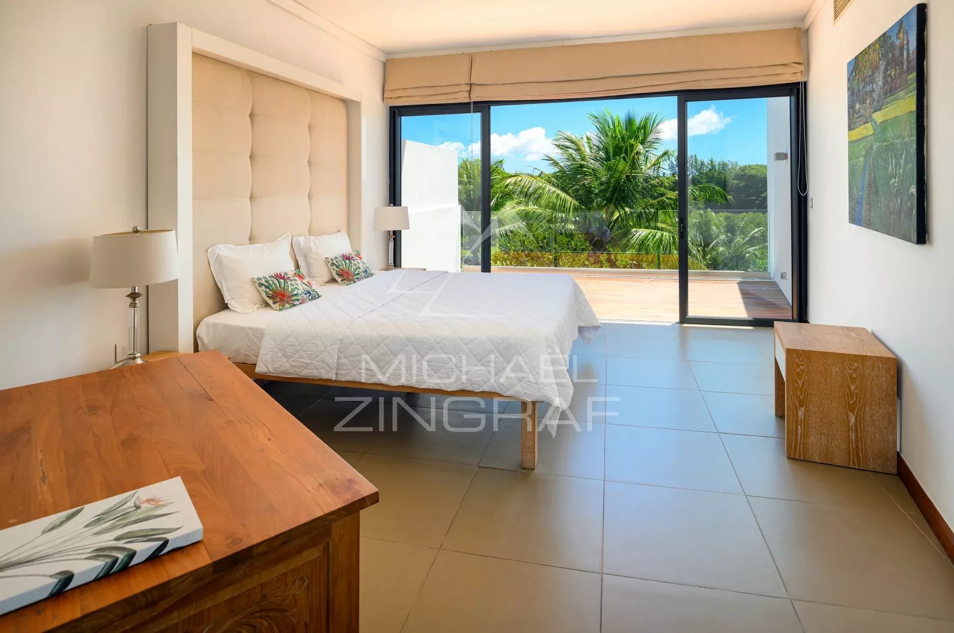 Hotel bedroom with a king bed, tufted headboard, nightstands, and sliding glass doors to a balcony overlooking palm trees and blue sky.