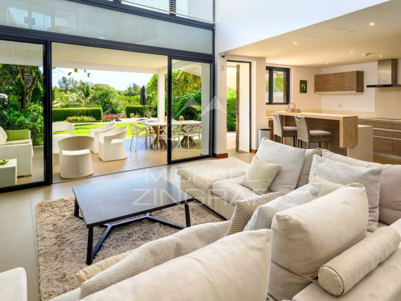 Open-plan living room with beige sofas and a wooden coffee table, sliding glass doors to a sunny outdoor patio rich with greenery.