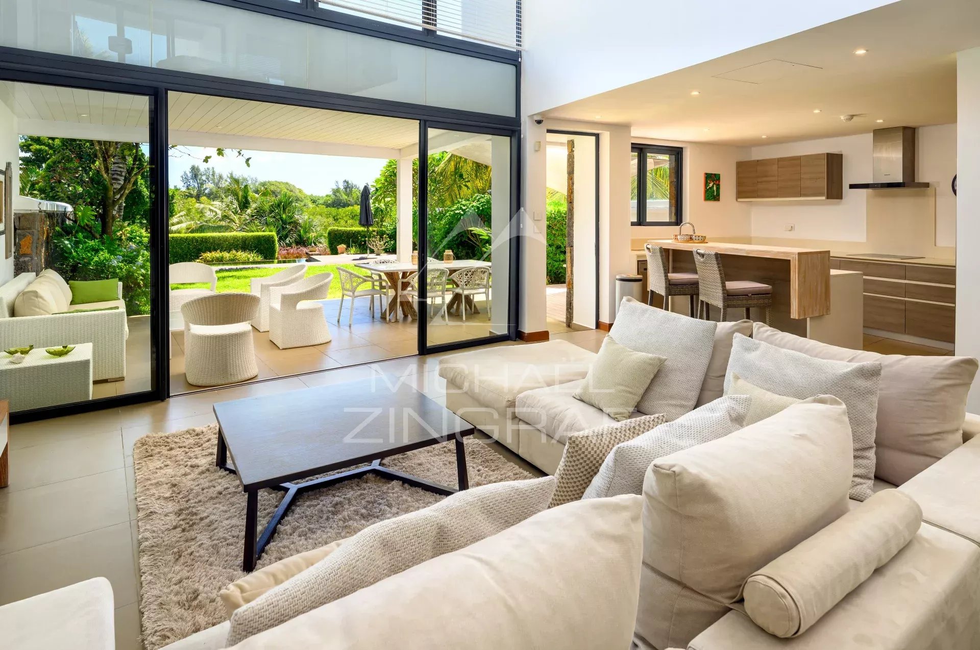 Open-plan living room with beige sofas and a wooden coffee table, sliding glass doors to a sunny outdoor patio rich with greenery.