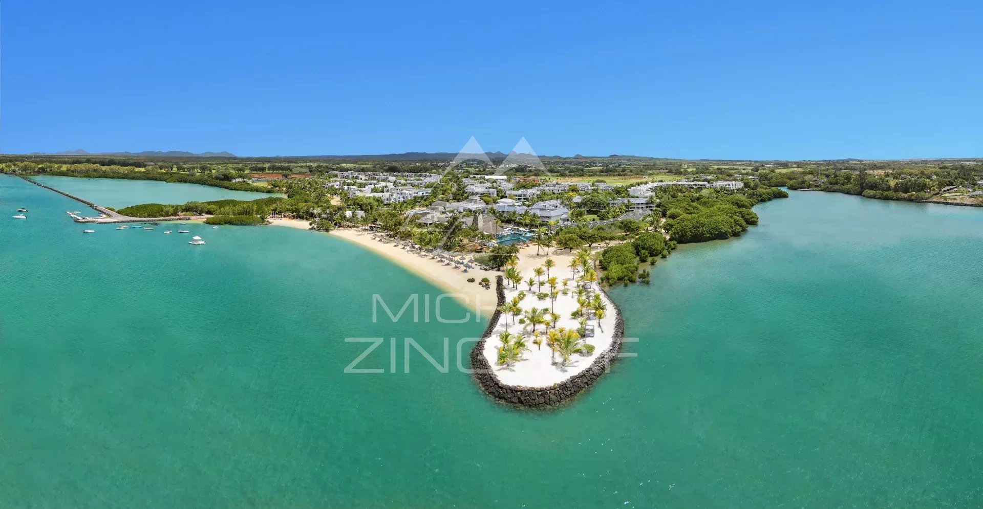 Aerial view of a tropical resort on a curved sandy peninsula with palm trees, turquoise water, and white buildings in the background.