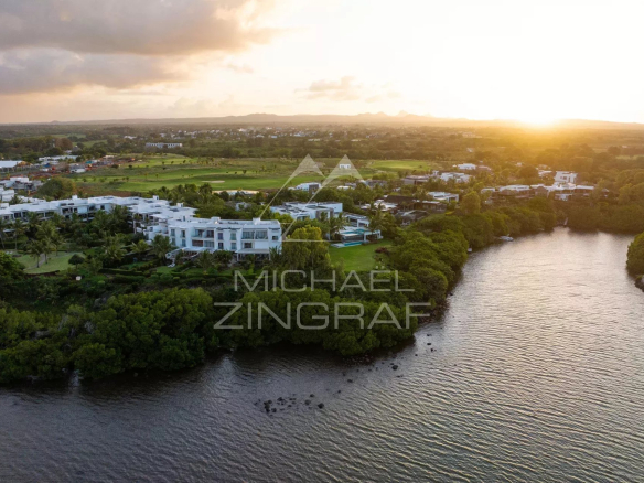 Aerial view of white modern villas along a curved river at sunset, with green landscaping and a distant golf course.