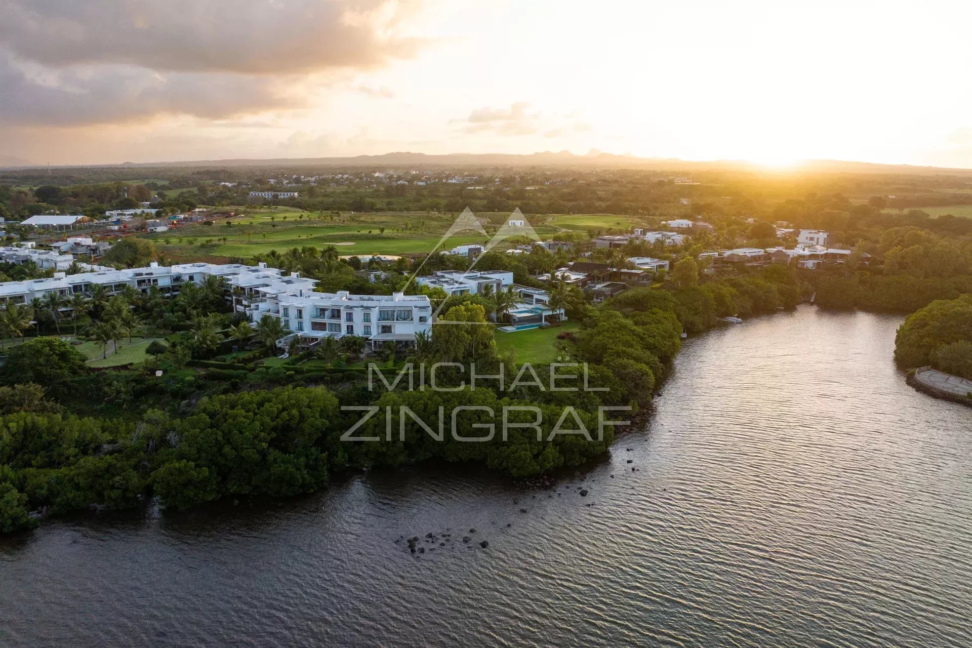 Aerial view of white modern villas along a curved river at sunset, with green landscaping and a distant golf course.