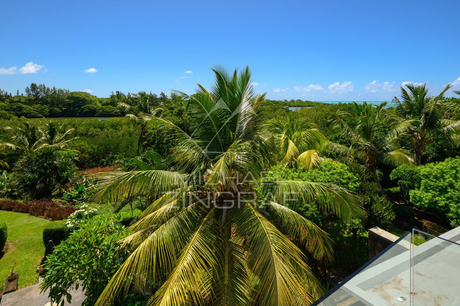 Tropical garden with tall palm trees, dense green foliage, and a bright blue sky; distant water visible on the horizon, with a glass railing in the lower right corner.