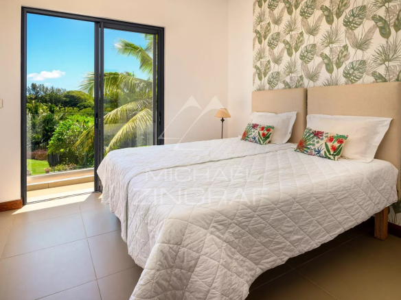 Bright bedroom with a white quilted bed, tropical wallpaper, and a sliding glass door to a balcony overlooking greenery.