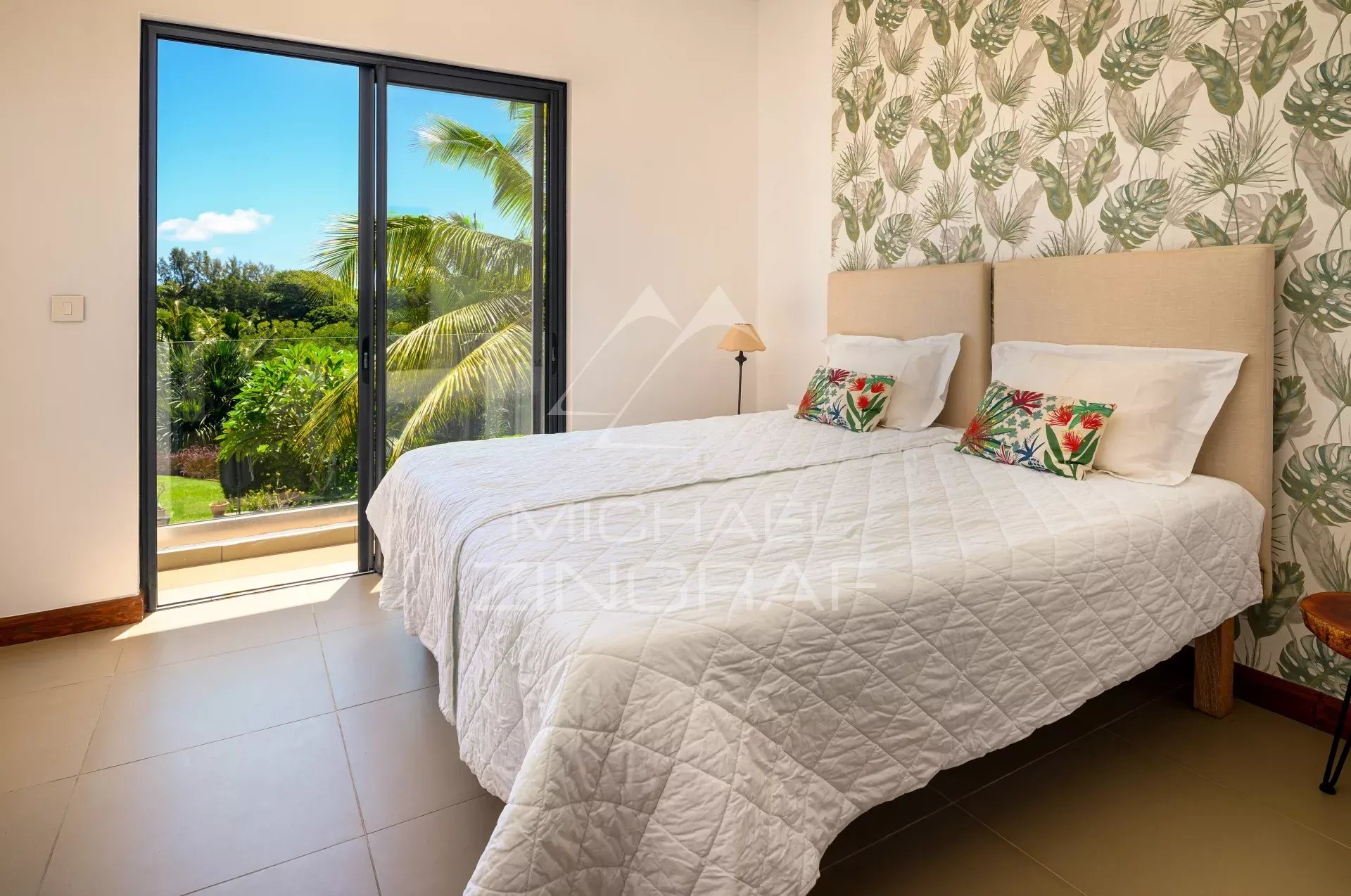 Bright bedroom with a white quilted bed, tropical wallpaper, and a sliding glass door to a balcony overlooking greenery.