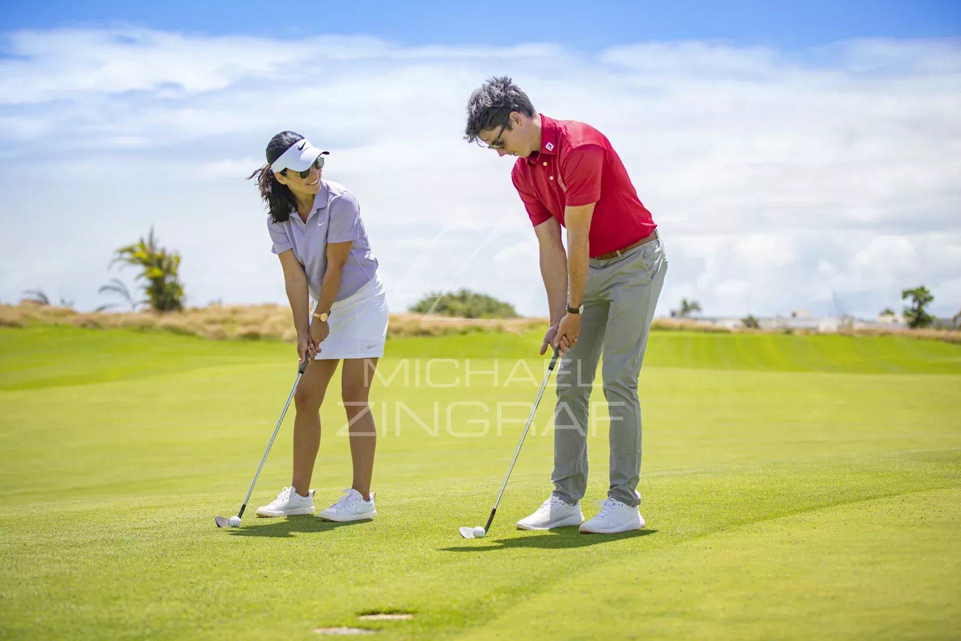 A man and a woman on a sunny golf course, each with a putter, bending over to line up a putt together.