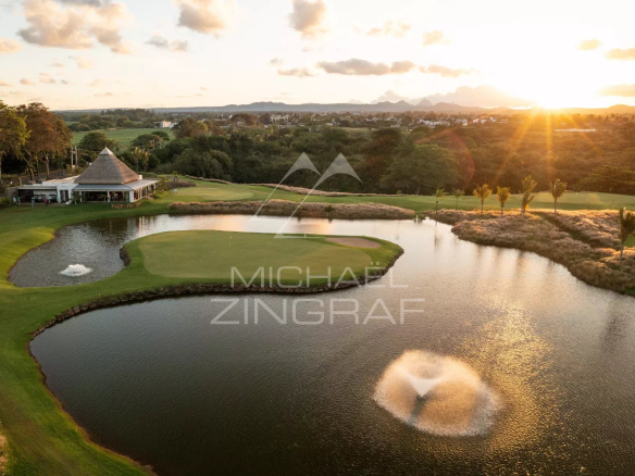 Aerial view of a golf course at sunset with a green island, surrounding water hazards, a fountain on the pond, and a thatched-roof clubhouse nearby.