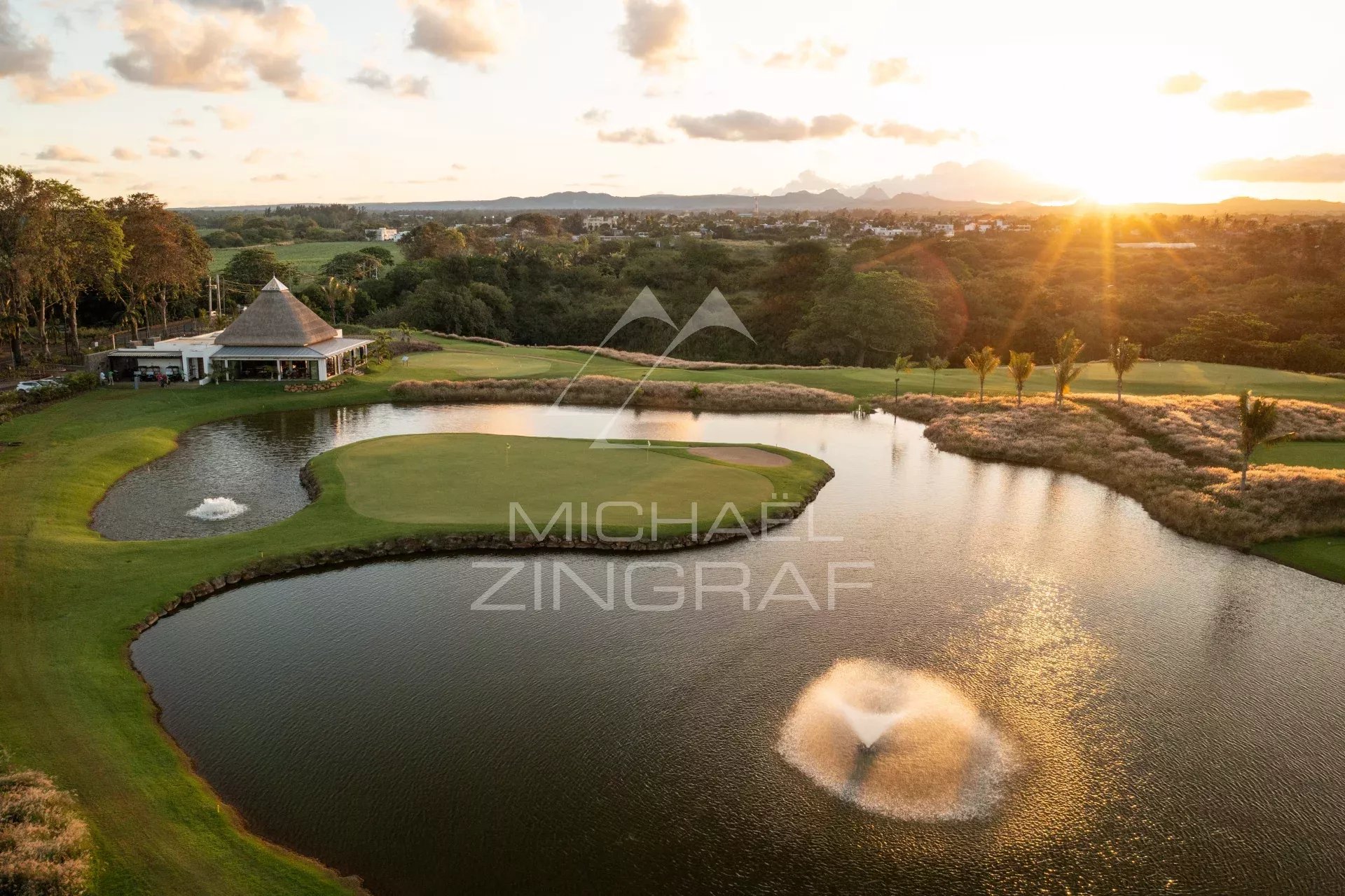 Aerial view of a golf course at sunset with a green island, surrounding water hazards, a fountain on the pond, and a thatched-roof clubhouse nearby.
