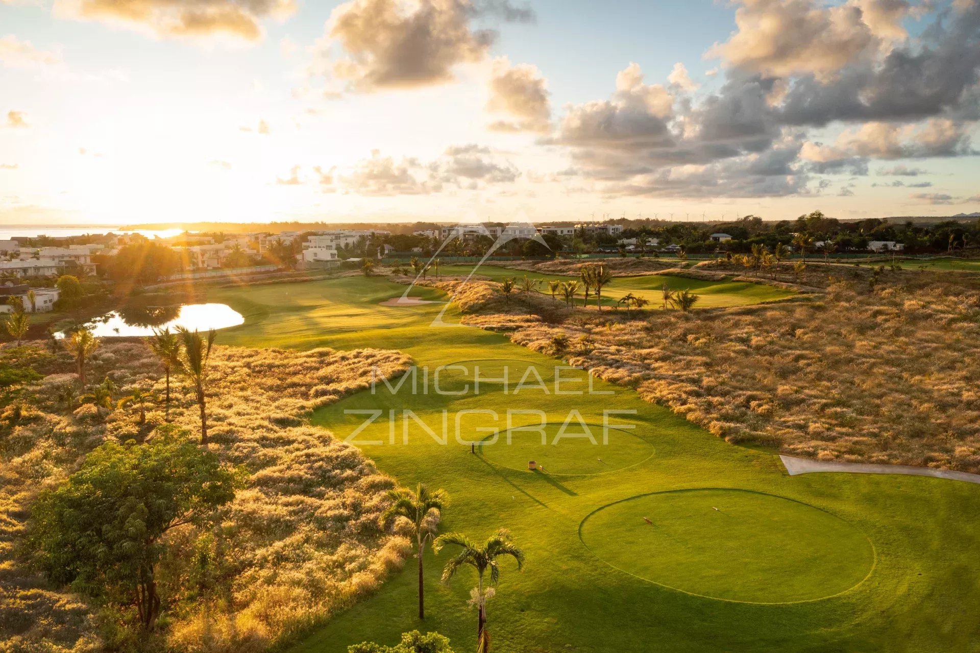 Aerial view of a golf course at sunset with green fairways, sand bunkers, a small pond, and nearby buildings in the distance.