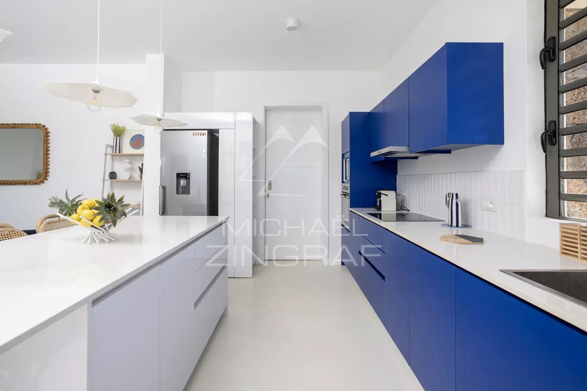 Bright modern kitchen with a white island, blue cabinets, a stainless fridge, and a fruit bowl on the counter.