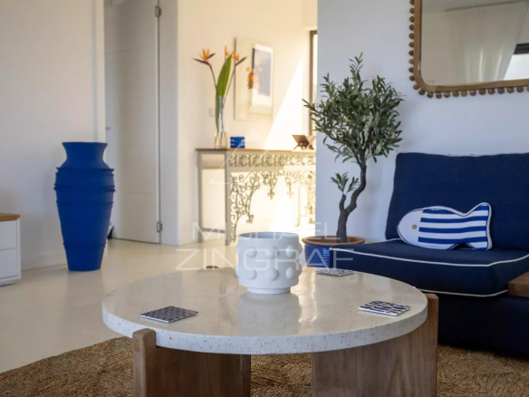Bright living room with a round speckled coffee table, navy sofa, and a striped bolster pillow. A tall blue vase and a potted plant sit nearby by a decorative mantel and mirror.