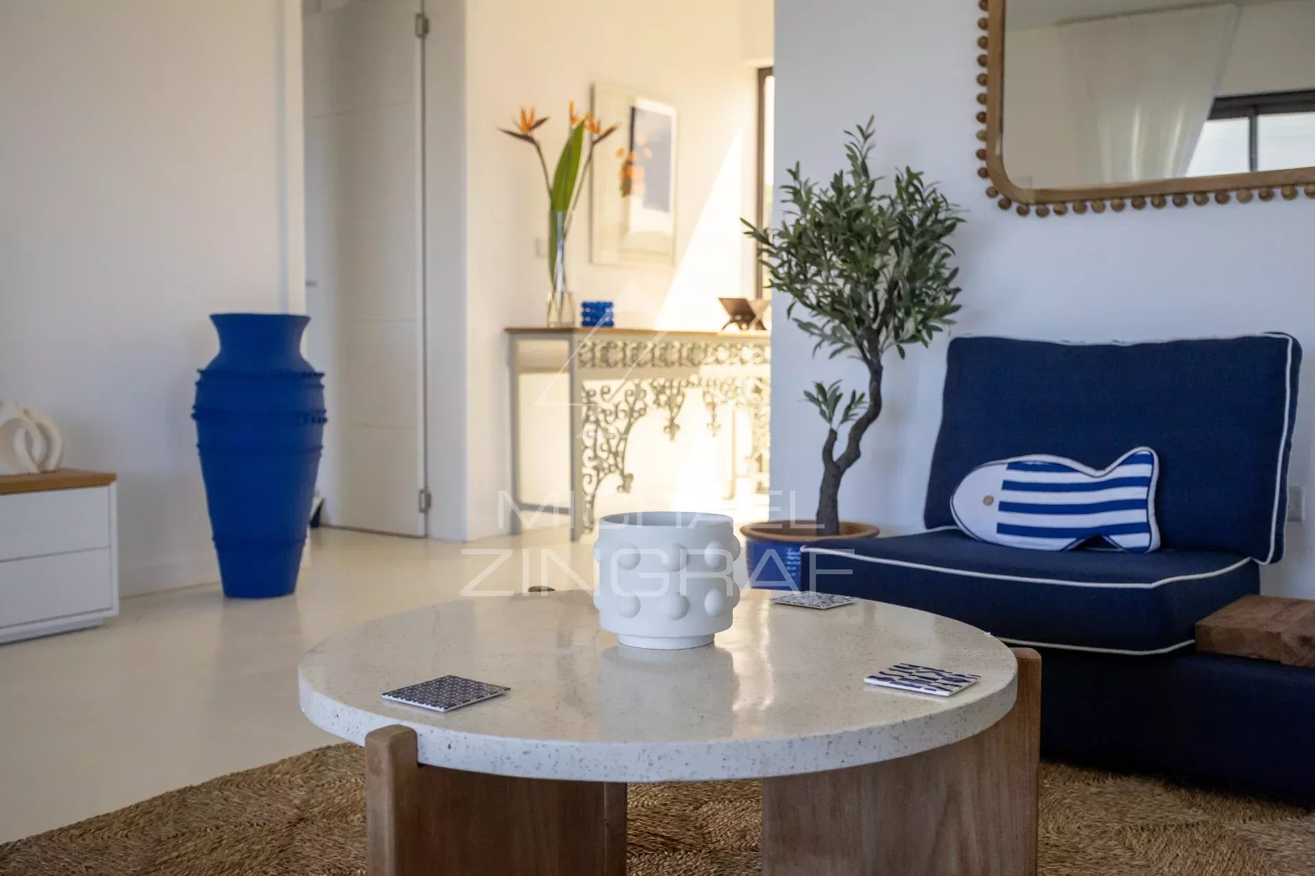 Bright living room with a round speckled coffee table, navy sofa, and a striped bolster pillow. A tall blue vase and a potted plant sit nearby by a decorative mantel and mirror.