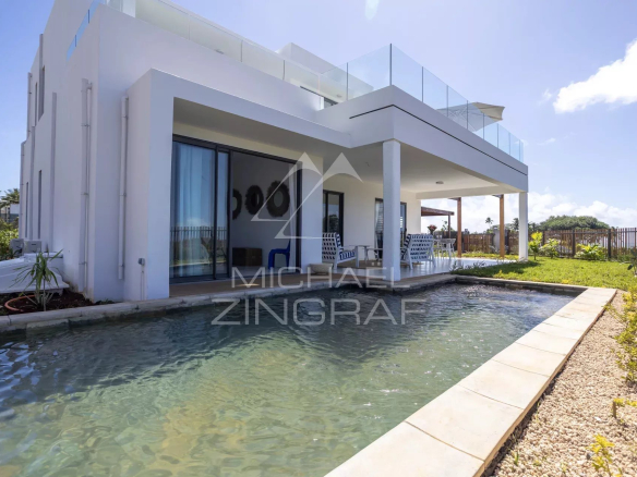 Modern white two-story house with a rectangular pool and a covered patio with blue and white seating under a sunny sky.