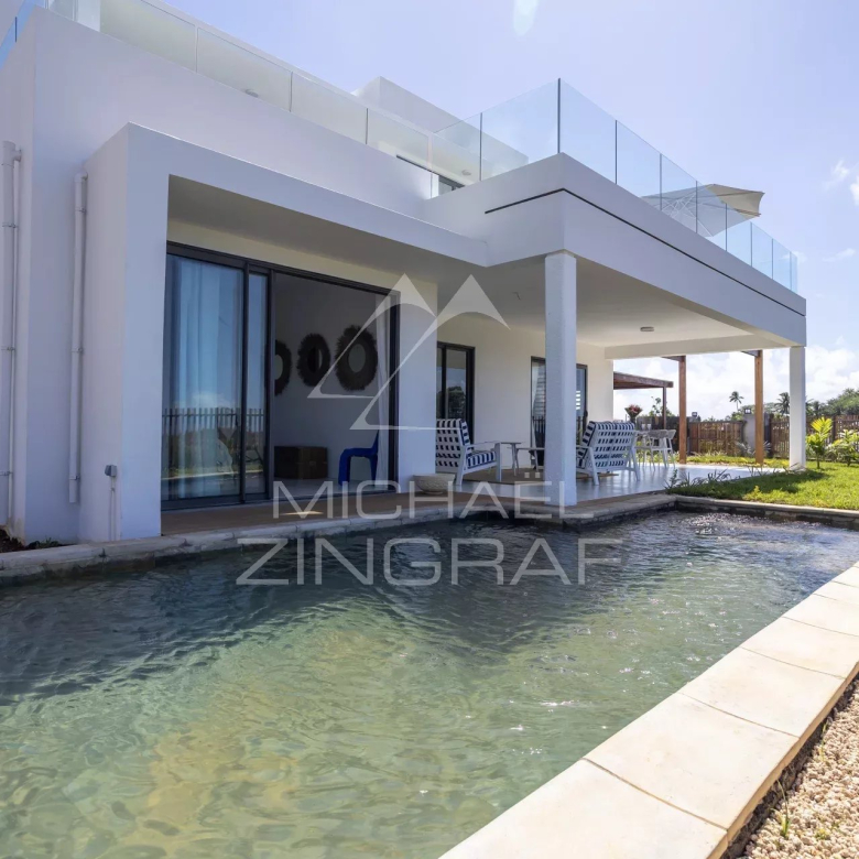 Modern white two-story house with a rectangular pool and a covered patio with blue and white seating under a sunny sky.