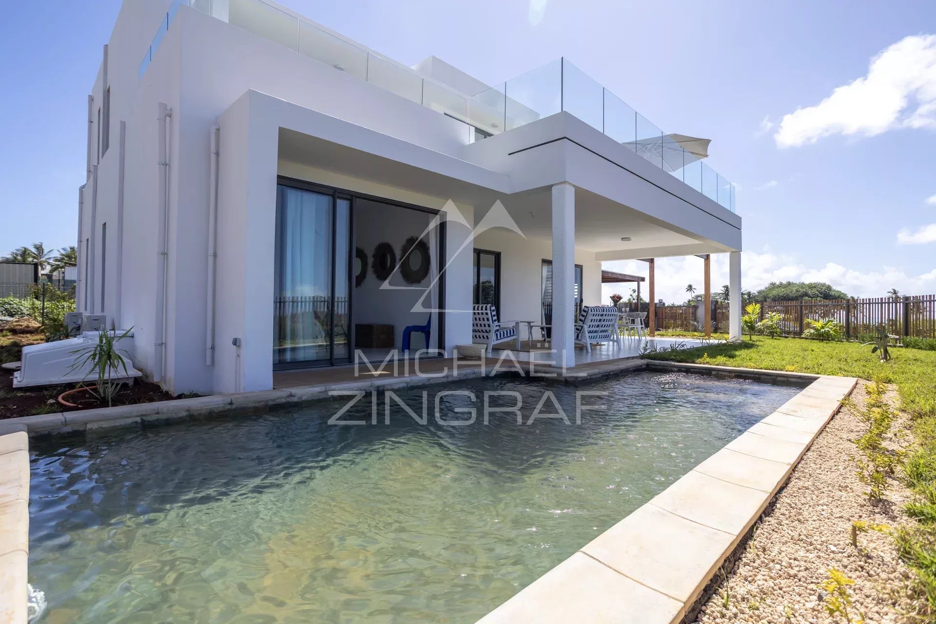 Modern white two-story house with a rectangular pool and a covered patio with blue and white seating under a sunny sky.