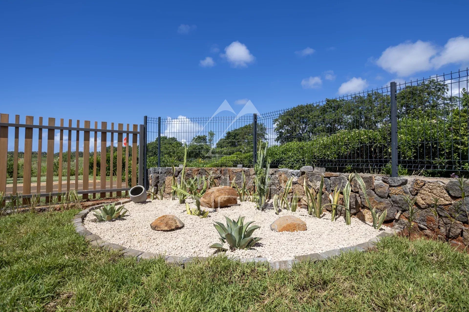 Rock garden with gravel, succulents, and large decorative stones behind a metal and wooden fence under a blue sky.