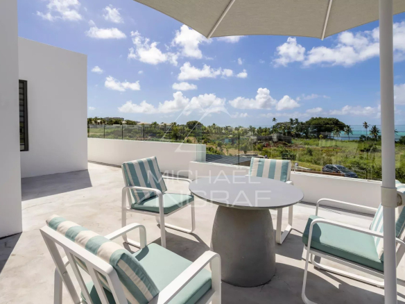 Outdoor patio on a sunny balcony with a round table and striped turquoise cushions, overlooking a coastal landscape with palm trees and the ocean behind a glass railing.