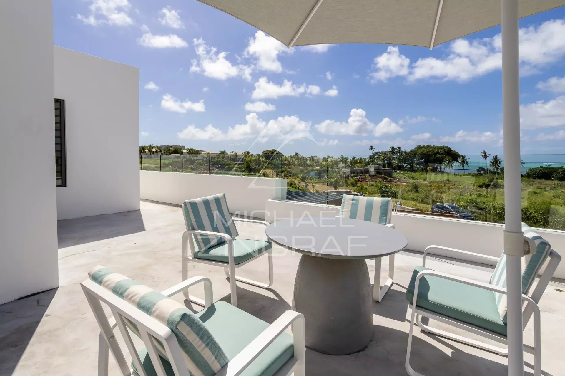 Outdoor patio on a sunny balcony with a round table and striped turquoise cushions, overlooking a coastal landscape with palm trees and the ocean behind a glass railing.