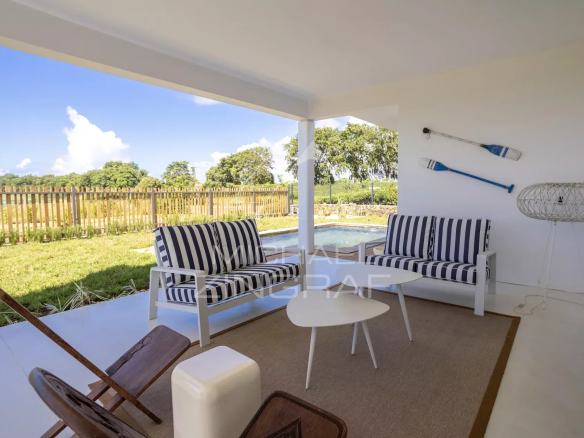 Covered outdoor seating area with white furniture and navy striped cushions, overlooking a pool and garden under a blue sky.