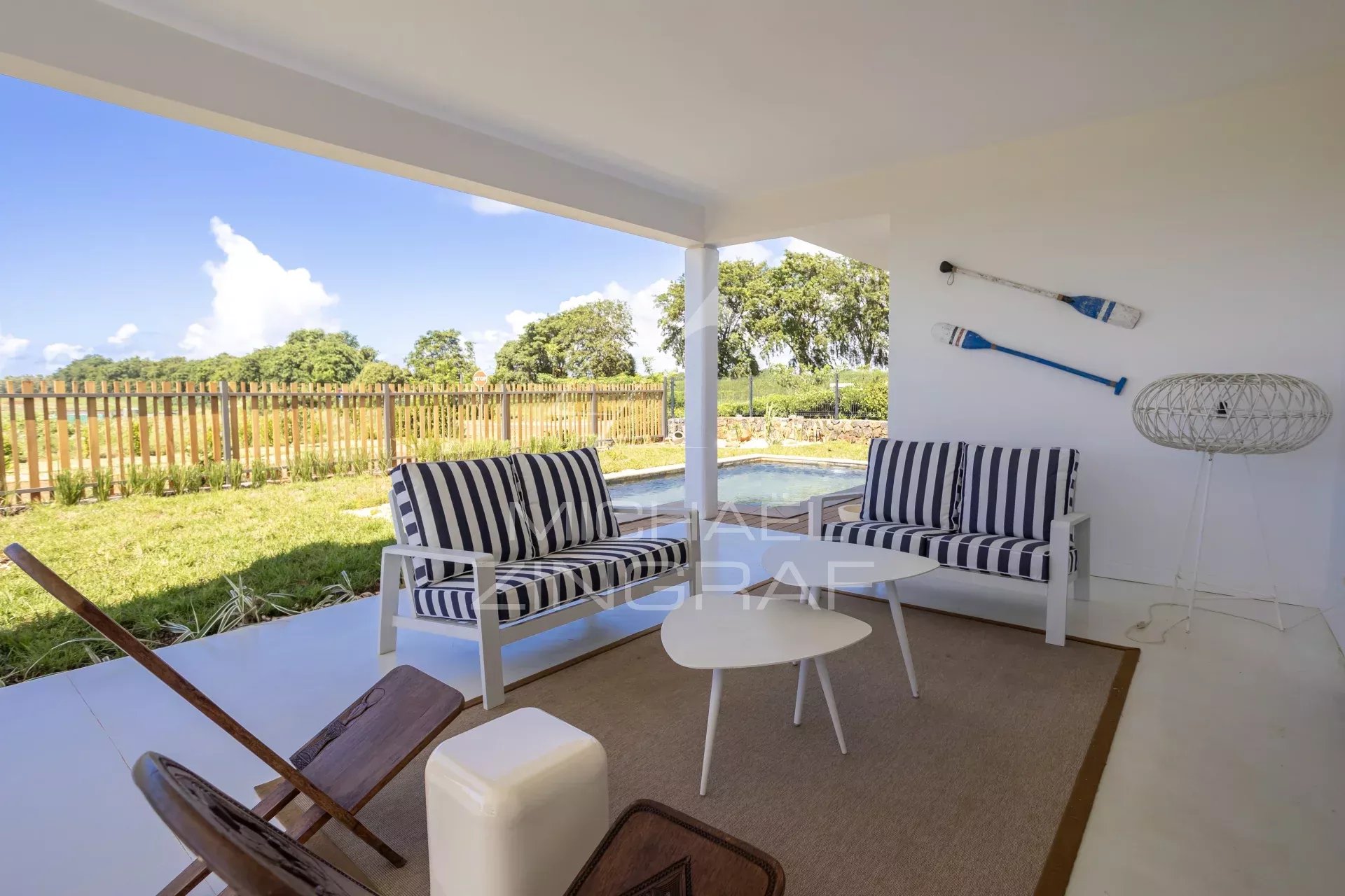 Covered outdoor seating area with white furniture and navy striped cushions, overlooking a pool and garden under a blue sky.