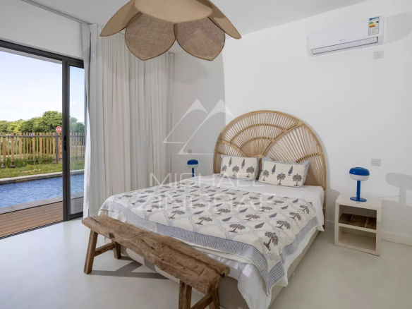 Bright coastal bedroom with a woven rattan headboard, white walls, and a sliding glass door opening to a pool area outside.