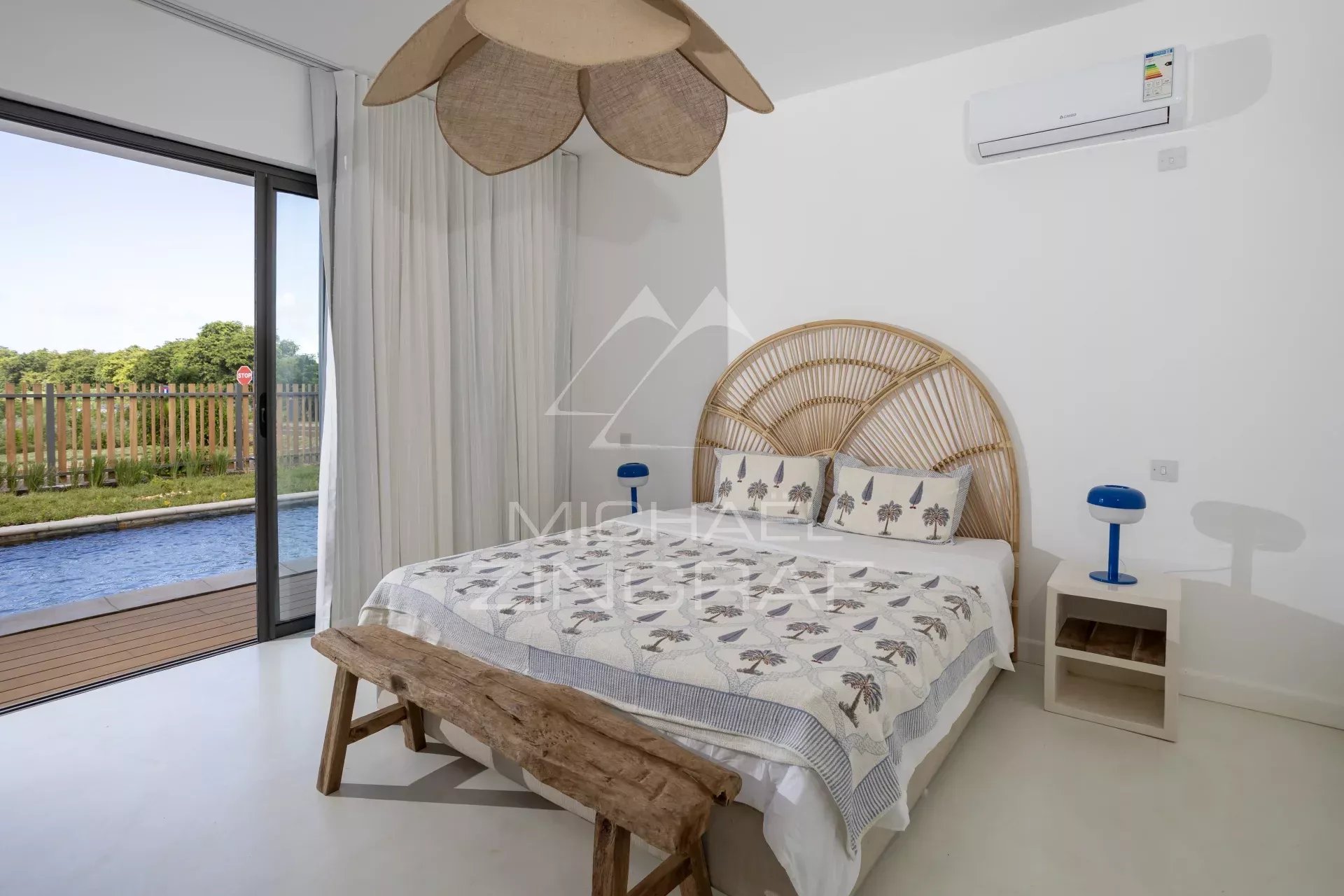 Bright coastal bedroom with a woven rattan headboard, white walls, and a sliding glass door opening to a pool area outside.