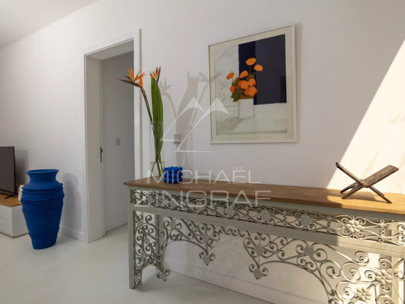 Living room entry area with a decorative metal console table, a wooden top, and a tall glass vase holding orange flowers; a framed abstract painting above and a wooden sculptural piece on the right side of the table.