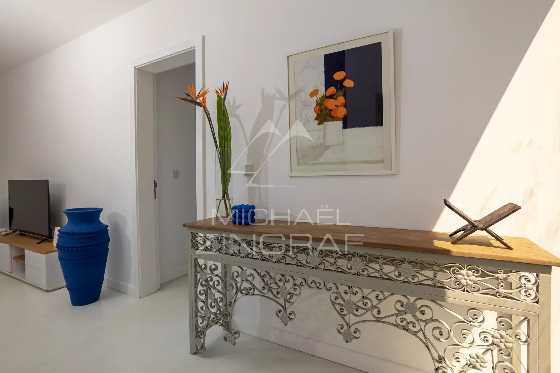 Living room entry area with a decorative metal console table, a wooden top, and a tall glass vase holding orange flowers; a framed abstract painting above and a wooden sculptural piece on the right side of the table.