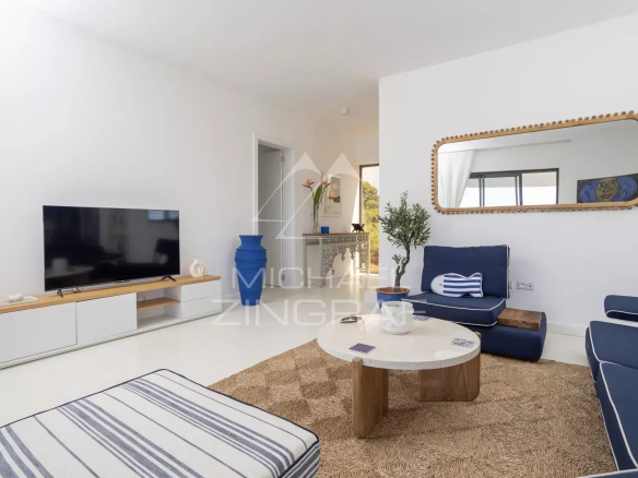 Bright living room with white walls, navy blue sofas, a round light-colored coffee table on a woven rug, and a large oval mirror with a beaded wooden frame above the seating area.