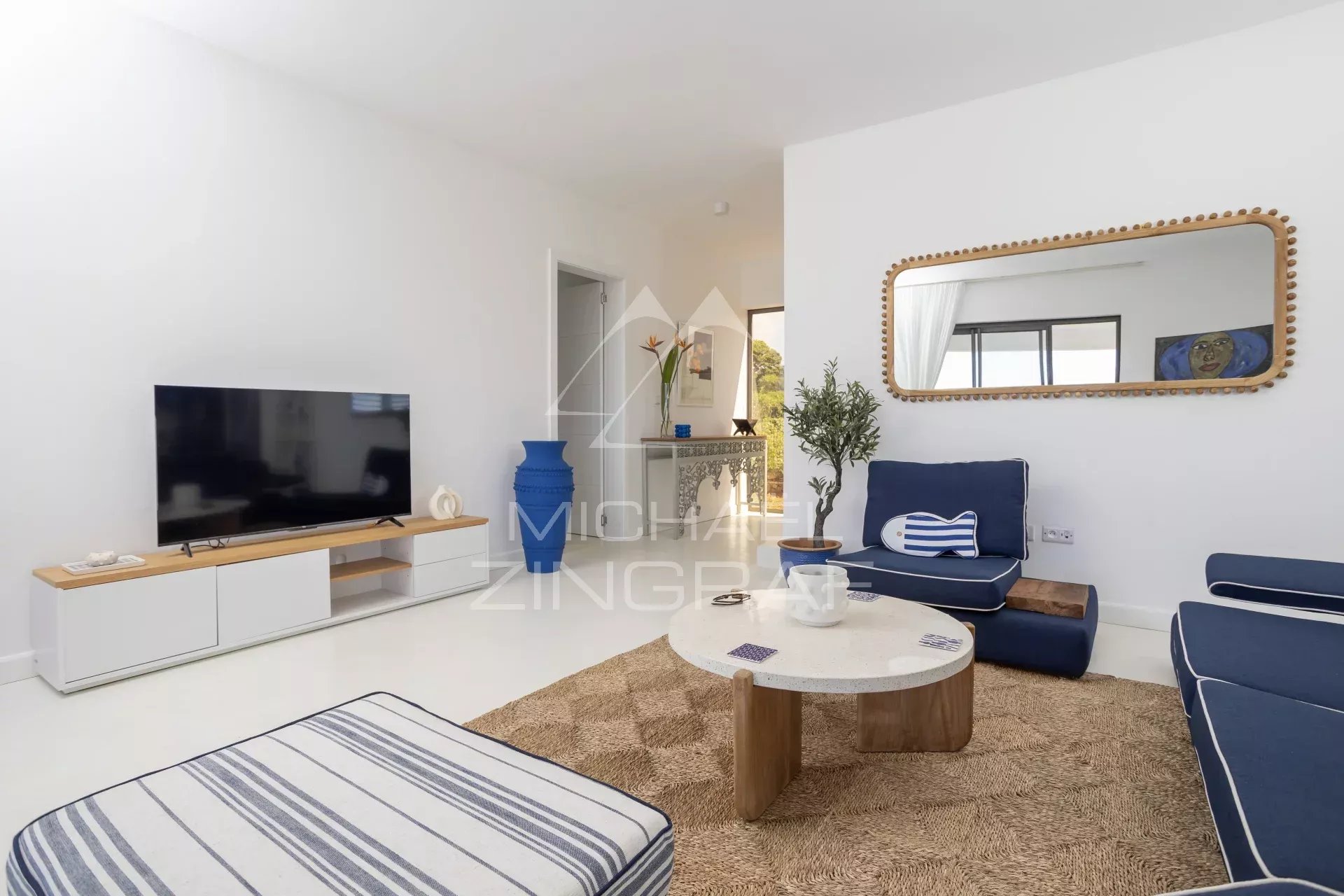 Bright living room with white walls, navy blue sofas, a round light-colored coffee table on a woven rug, and a large oval mirror with a beaded wooden frame above the seating area.