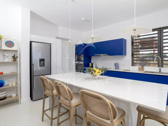 Contemporary white kitchen with blue cabinets, a long white island, and wooden chairs around it; stainless fridge and large window on the right.