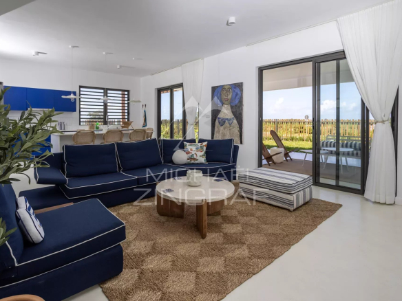 Bright coastal living room with navy sofas, round wooden coffee table, and a woven jute rug, opening to a sunny patio through glass doors.