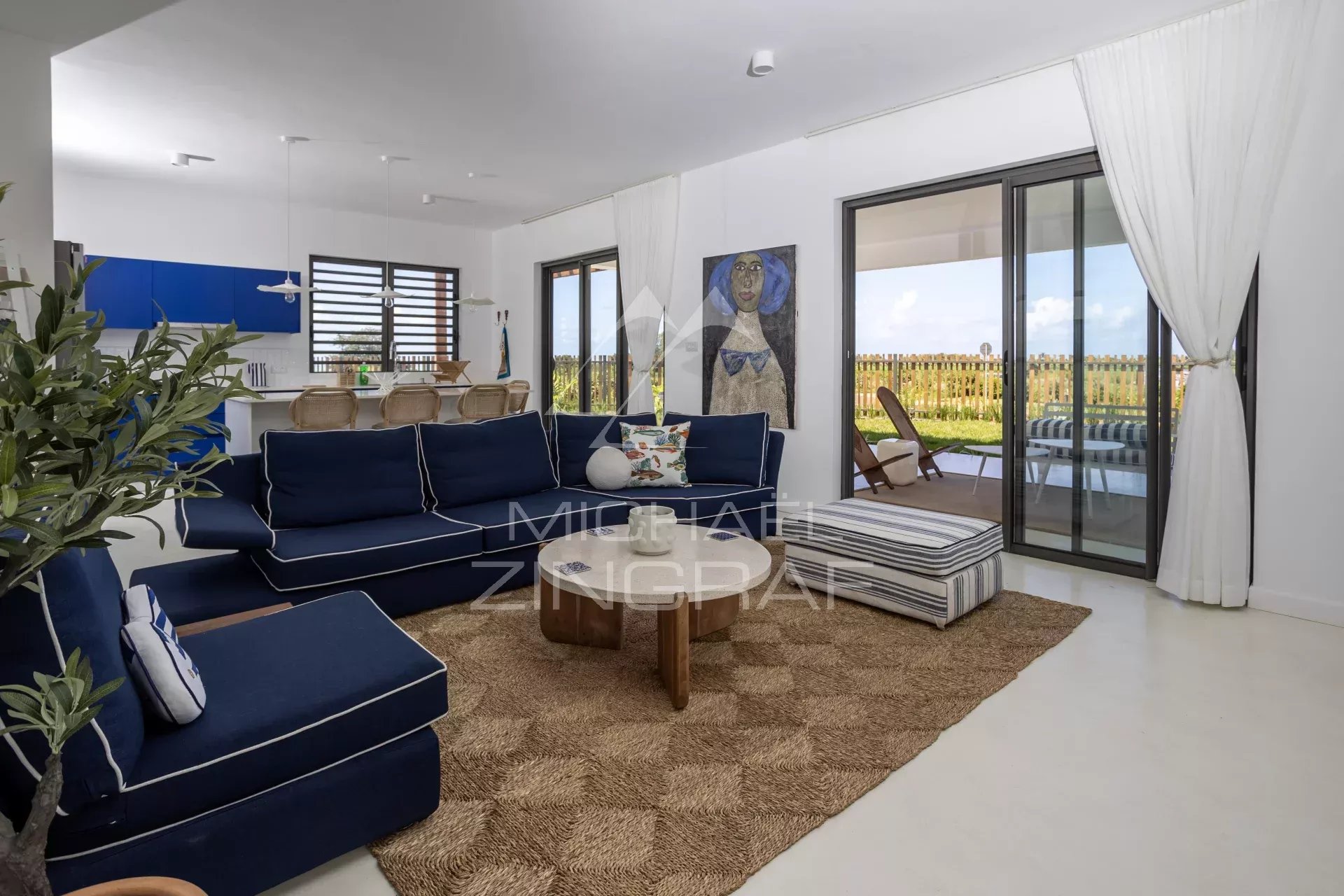 Bright coastal living room with navy sofas, round wooden coffee table, and a woven jute rug, opening to a sunny patio through glass doors.
