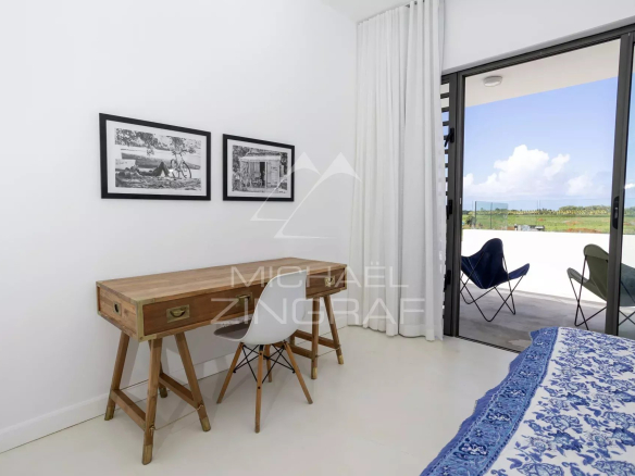 Bedroom with a wooden desk and white chair beside two black-and-white framed photos on white walls, next to a sliding glass door to a balcony with outdoor chairs.