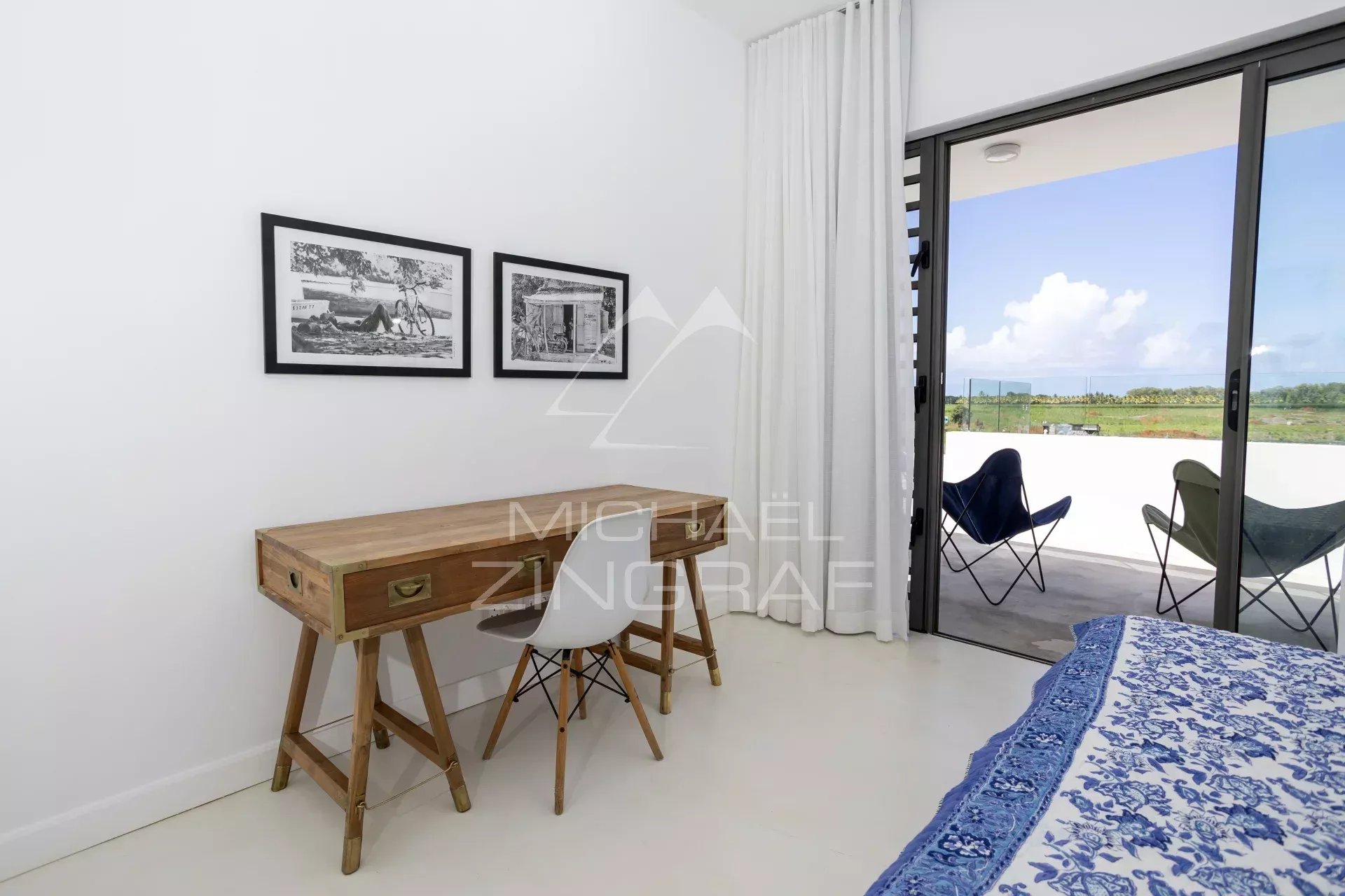 Bedroom with a wooden desk and white chair beside two black-and-white framed photos on white walls, next to a sliding glass door to a balcony with outdoor chairs.