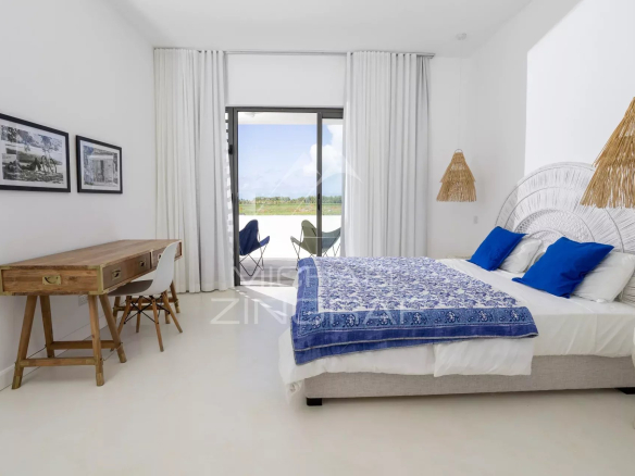 Bright bedroom with a white bed, blue pillows and a blue patterned throw; a wooden desk and balcony doors in view.