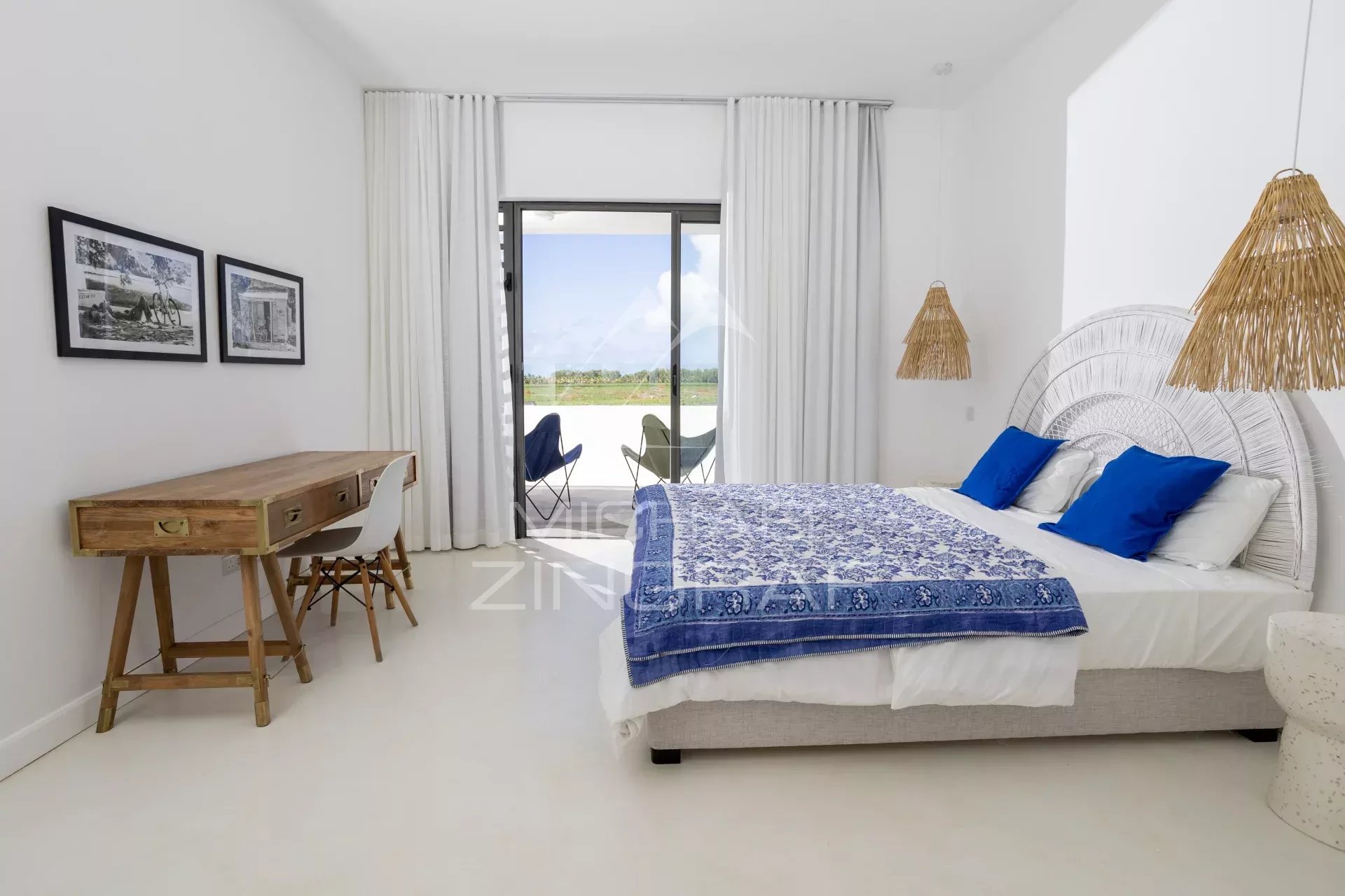 Bright bedroom with a white bed, blue pillows and a blue patterned throw; a wooden desk and balcony doors in view.